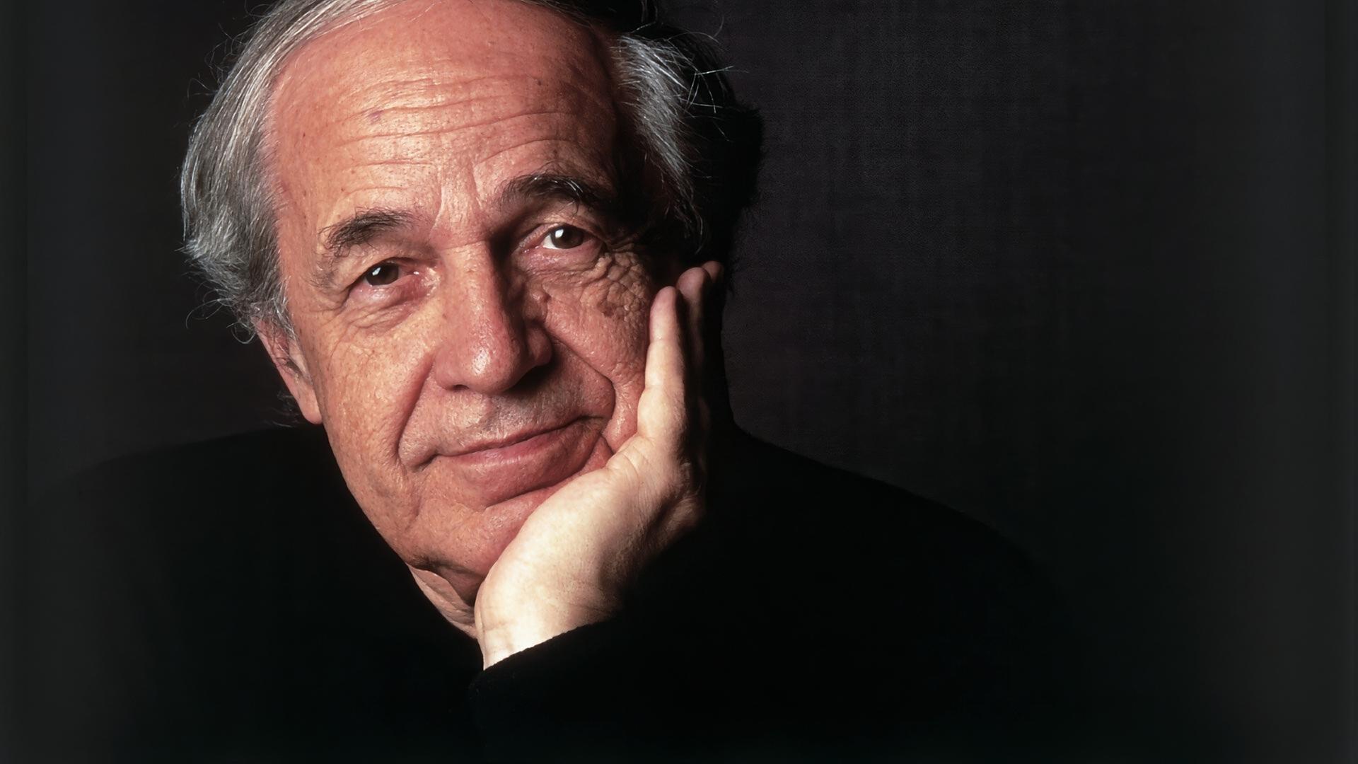 Das Bild zeigt einen älteren Mann mit einem nachdenklichen Ausdruck. Er hat graue, leicht gewellte Haare, die an den Seiten kürzer geschnitten sind, und eine helle Hautfarbe. Der Mann hat einen leichten Bartschatten und trägt ein dunkles, schlichtes Oberteil. Er stützt sein Kinn mit der linken Hand, während seine rechte Hand auf der Tischkante liegt. Der Hintergrund des Bildes ist dunkel und einheitlich, was den Fokus auf das Gesicht des Mannes lenkt. Die Lichtsetzung betont seine Gesichtszüge und verleiht dem Bild eine subtile Wärme.