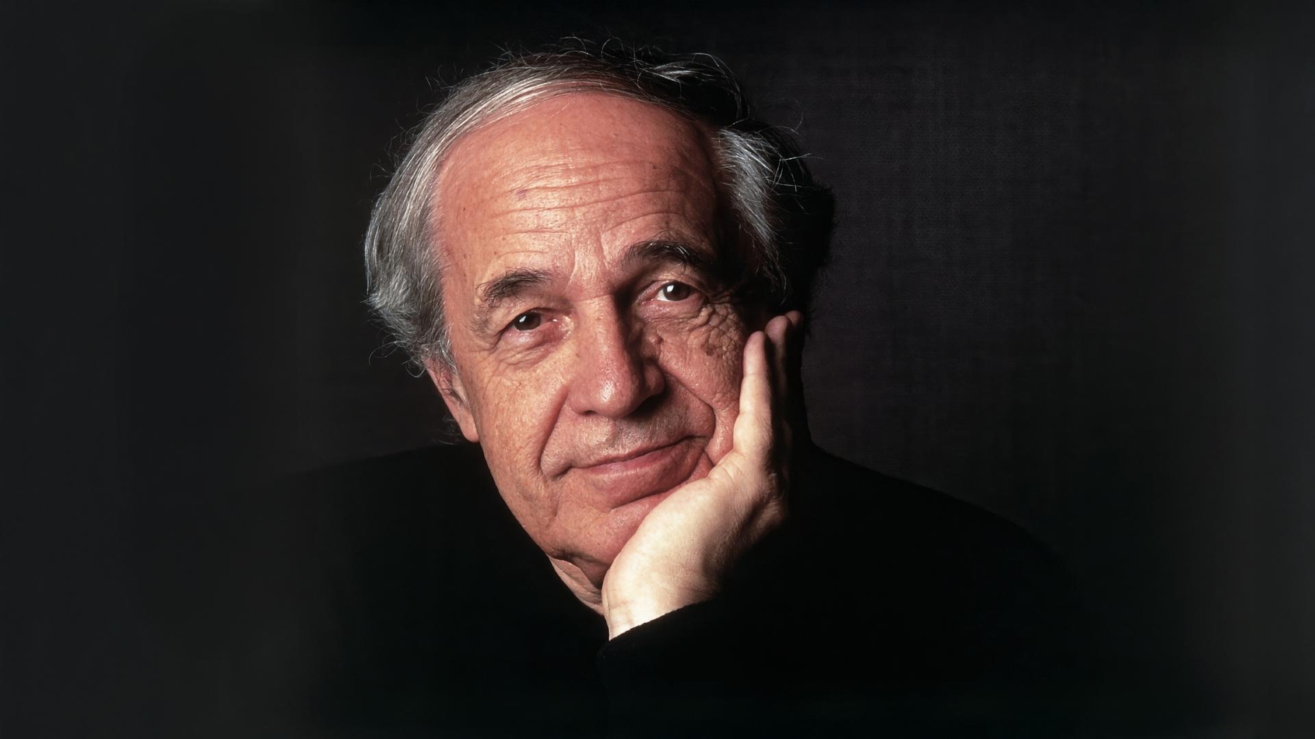 Das Bild zeigt einen älteren Mann mit einem nachdenklichen Ausdruck. Er hat graue, leicht gewellte Haare, die an den Seiten kürzer geschnitten sind, und eine helle Hautfarbe. Der Mann hat einen leichten Bartschatten und trägt ein dunkles, schlichtes Oberteil. Er stützt sein Kinn mit der linken Hand, während seine rechte Hand auf der Tischkante liegt. Der Hintergrund des Bildes ist dunkel und einheitlich, was den Fokus auf das Gesicht des Mannes lenkt. Die Lichtsetzung betont seine Gesichtszüge und verleiht dem Bild eine subtile Wärme.