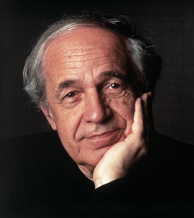 Das Bild zeigt einen älteren Mann mit einem nachdenklichen Ausdruck. Er hat graue, leicht gewellte Haare, die an den Seiten kürzer geschnitten sind, und eine helle Hautfarbe. Der Mann hat einen leichten Bartschatten und trägt ein dunkles, schlichtes Oberteil. Er stützt sein Kinn mit der linken Hand, während seine rechte Hand auf der Tischkante liegt. Der Hintergrund des Bildes ist dunkel und einheitlich, was den Fokus auf das Gesicht des Mannes lenkt. Die Lichtsetzung betont seine Gesichtszüge und verleiht dem Bild eine subtile Wärme.