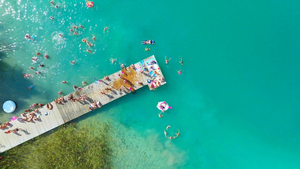 Das Bild zeigt eine Luftaufnahme des Wörthersees in Österreich, speziell während des Pink Lake Festivals. Im Vordergrund ist ein Holzsteg zu sehen, der ins Wasser führt. Auf dem Steg sind viele Menschen, die sich entspannen, liegen oder stehen. Einige haben Handtücher und bunte Strandmatten mitgebracht.   Im Wasser schwimmen zahlreiche Menschen, und es sind auch bunte Badeartikel wie aufblasbare Flöße zu erkennen. Die Wasseroberfläche hat einen klaren, türkisfarbenen Farbton, während am Ufer einige grüne Wasserpflanzen sichtbar sind. Die gesamte Szene strahlt eine festliche Atmosphäre aus, die mit Farben und dem geschäftigen Treiben der Festivalbesucher verbunden ist.   Die Umgebung wirkt sonnig und einladend, passend zu einem Sommerfestival, das der Vielfalt und Akzeptanz gewidmet ist.
