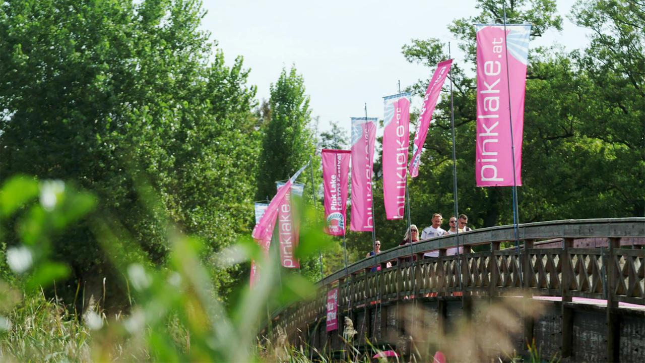Das Bild zeigt eine Holzbrücke, die durch eine parkähnliche Umgebung führt. Auf beiden Seiten der Brücke stehen hohe, grüne Bäume. Über die Brücke sind zahlreiche pinkfarbene Fahnen gespannt, die mit der Aufschrift "pinklake.at" versehen sind. Darunter sind mehrere Personen zu sehen, die entspannt entlang der Brücke gehen. Die Szene vermittelt eine fröhliche und einladende Atmosphäre, die die bunte und vielfältige Stimmung des Pink Lake Festivals widerspiegelt. Im Vordergrund sind auch einige grüne Pflanzen sichtbar, die dem Bild eine natürliche Note verleihen. Das Wetter scheint sonnig und klar zu sein.