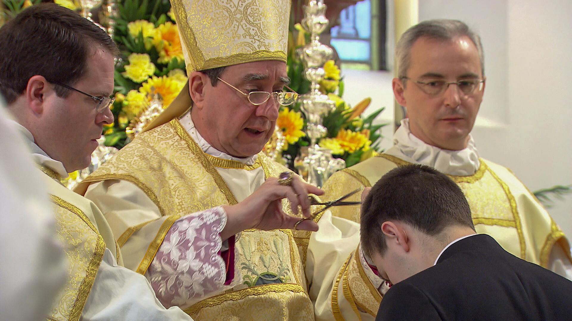 Das Bild zeigt eine Zeremonie in einem religiösen Kontext. Im Vordergrund ist ein junger Mann zu sehen, der sich leicht nach vorne beugt. Ein Bischof, gekleidet in eine prunkvolle, goldene liturgische Kleidung mit einem Mitra auf dem Kopf, schneidet mit einer Schere eine kleine Haarsträhne vom Kopf des jungen Mannes ab. Der Bischof hält die Schere in seiner rechten Hand, während er mit der linken Hand eine Handbewegung macht.  Im Hintergrund sind dekorative Blumenarrangements, überwiegend gelb, zu sehen. Neben dem Bischof stehen zwei andere Männer in ähnlicher, aber weniger aufwendiger liturgischer Kleidung, die aufmerksam auf die Zeremonie blicken. Die Atmosphäre wirkt feierlich und rituell.