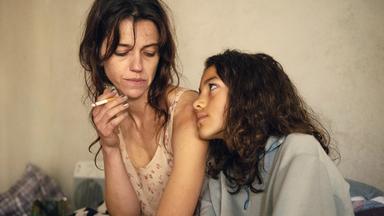 Filmszene: Eine Frau mit ungekämmten Haaren und Zigarette in der Hand lehnt neben einem jungen Mädchenund schaut auf dieses herab.