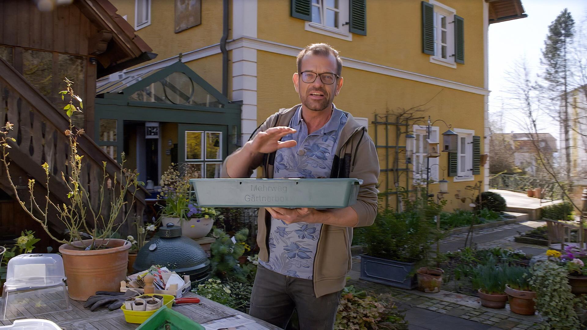 Ein Mann mit Brille und Bart steht vor einem gelben Haus mit grünem Fensterladen. Er hält eine grüne Pflanzenschale in den Händen und spricht.