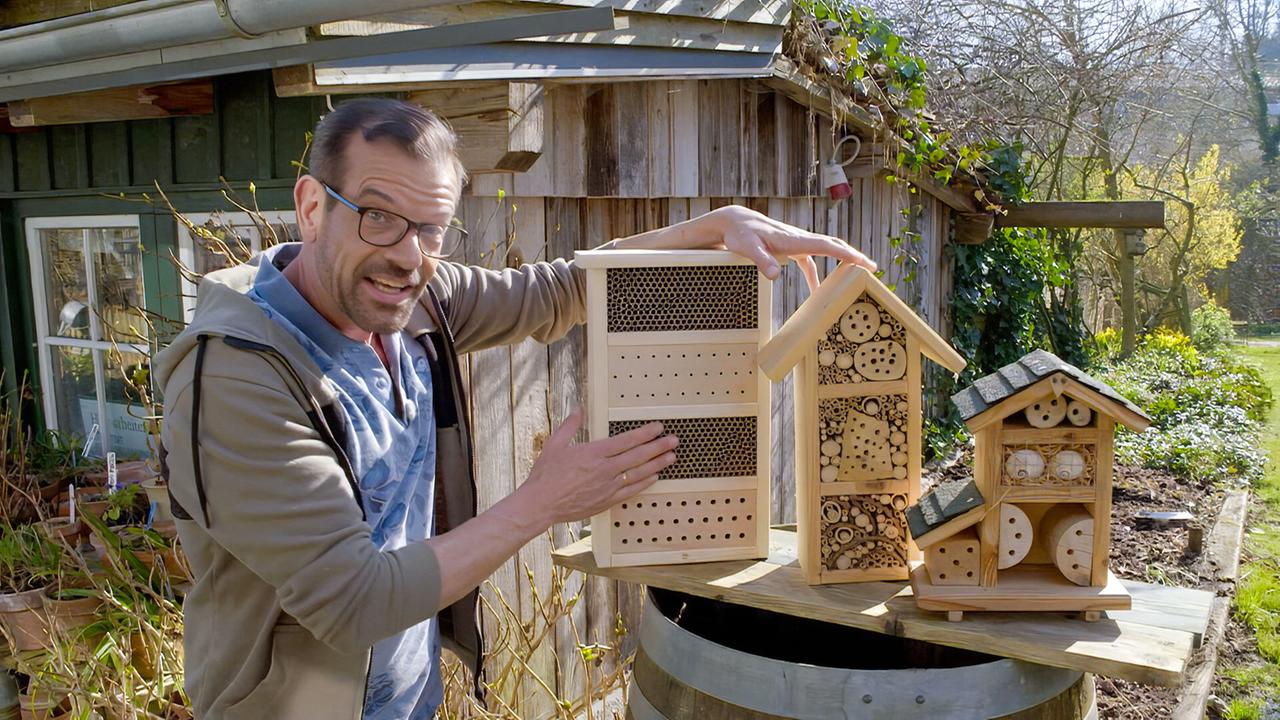 Ein Mann mit Brille und Bart steht im Garten vor einer Holzhütte und zeigt auf mehrere Insektenhotels aus Holz.