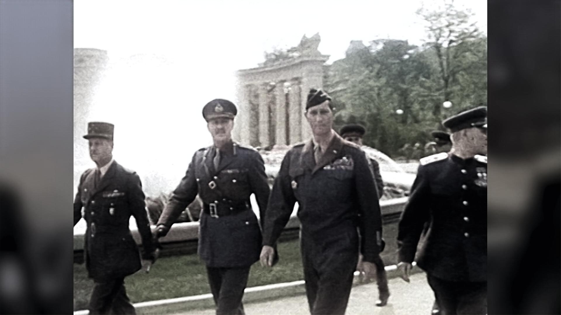 Das Bild zeigt eine Gruppe von vier Soldaten, die in militärischer Uniform gehen. Sie befinden sich auf einem Platz, der von einem Brunnen umgeben ist, und im Hintergrund sind eine Reihe von Säulen oder Gebäuden zu sehen, die auf eine architektonische Struktur hindeuten. Die Soldaten tragen unterschiedliche Uniformen, die auf verschiedene Ränge und Nationalitäten hinweisen könnten. Die Szene wirkt historisch, vermutlich aus der Zeit nach dem Zweiten Weltkrieg. Die allgemeine Atmosphäre vermittelt den Eindruck eines offiziellen Anlasses oder einer Zeremonie. Die Bildqualität ist in Schwarz-Weiß, was den zeitlichen Kontext verstärkt. Es gibt auch einige Bäume und Laternen im Hintergrund, die zur Umgebung beitragen.