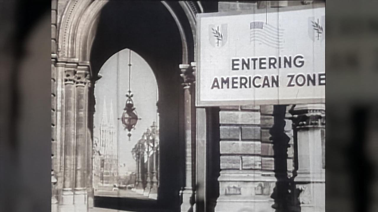 Das Bild zeigt einen historischen Standort im amerikanischen Sektor in Wien. Der Blick ist auf einen großen Torbogen gerichtet, in dem sich ein Schild mit der Aufschrift „ENTERING AMERICAN ZONE“ befindet. Das Schild ist groß und deutlich lesbar, mit schwarzen Buchstaben auf weißem Grund. Neben dem Schild sind verschiedene Symbole und Flaggen abgebildet.  Im Hintergrund ist ein Teil der städtischen Architektur sichtbar, darunter dekorative Elemente und historische Gebäude. Es sind auch Laternen hängend zu sehen, die dem Bild eine historische Atmosphäre verleihen. Der Stil der Architektur deutet auf eine eher prunkvolle Gestaltung hin, typisch für das Wien der damaligen Zeit. Die gesamte Szene vermittelt ein Gefühl des Wandels und der Öffnung, das nach dem Ende des Zweiten Weltkriegs in Wien stattfand.
