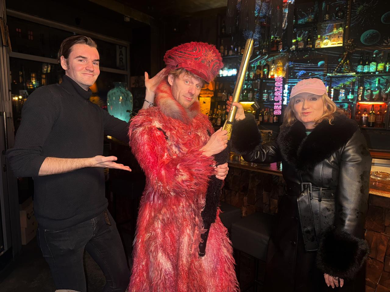 Drei Menschen an einer Bar. Im Mittelpunkt ein extravagant gekleideter Mann in rotem Pelzmantel mit Hut.