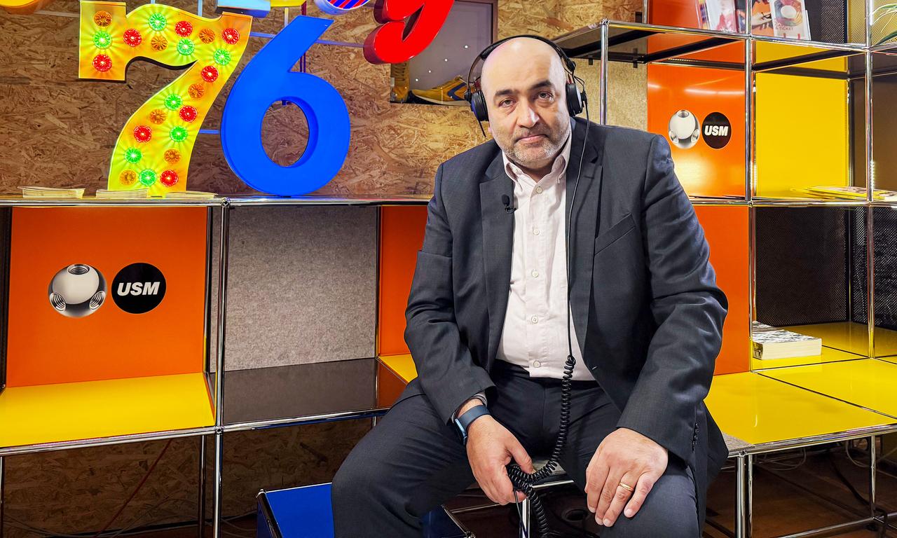 Omid Nouripour trägt Kopfhörer und sitzt im Café Frankfurter Massif Central vor einer Wand mit Leuchtbuchstaben. Darauf steht: Guilty 76.