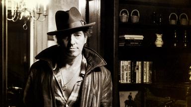 Bruce Springsteen steht mit Hut und Lederjacke in einenm Haus vor zwei Bücherregalen. Das Bild ist schwaz-weiß.