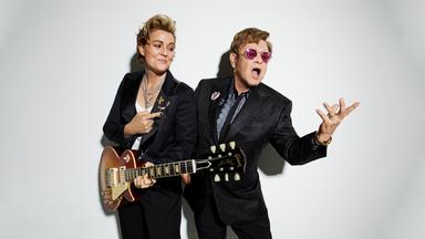 Brandi Carlile steht mit umgehängter Gitarre neben Elton John und schaut ihn an und zeigt auf ihn. Elton John trägt einen glitzernen Anzug und rosa Brille und singt.
