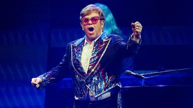 Elton John steht in glitzernem Anzug und mit roter Brille auf der Bühne und singt.