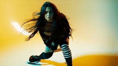 Vor einem gelben Hintergrund ist Amy Lee von Evanescence die mitten in einer schwungvollen Bewegung fotografiert wird. Ihre Haare fliegen durch die Luft und sie blickt in die Kamera