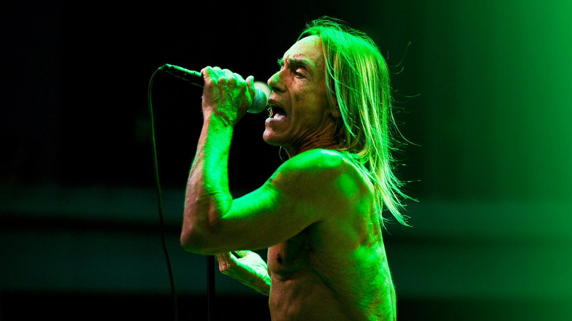 Iggy Pop steht oberkörperfrei auf der Bühne und singt ins Mikrofon.