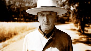 Marius Müller-Westernhagen trägt einen Hut, steht auf einem Feldweg und blickt in die Kamera.
