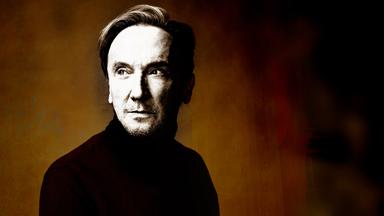 Marius Müller-Westernhagen sitzt in schwarzem Rollkragenpulli vor einer braunen Wand und schaut leicht nach rechts.