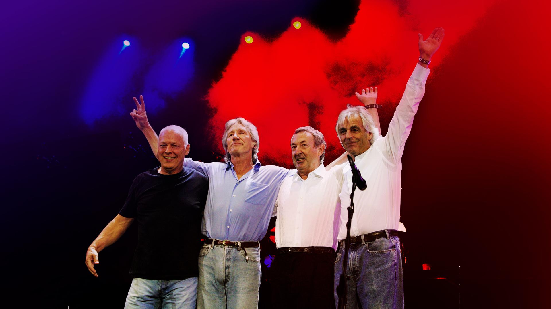 Vier Bandmitglieder von Pink Floyd stehen Arm in Arm auf einer Bühne.