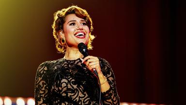 Raye steht in schwarzem Glitzerkleid auf der Bühne und singt lächelnd ins Mikrofon in ihrer Hand.