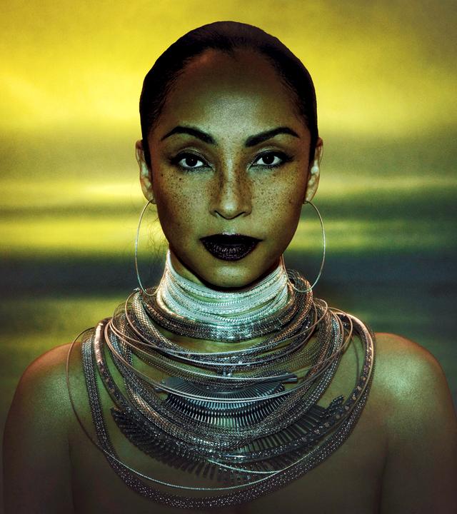 Sade Adu trägt große Kreolen und ganz viele Ketten um den Hals. Ihre Haare sind streng zurückgebunden und sie blickt in die Kamera.
