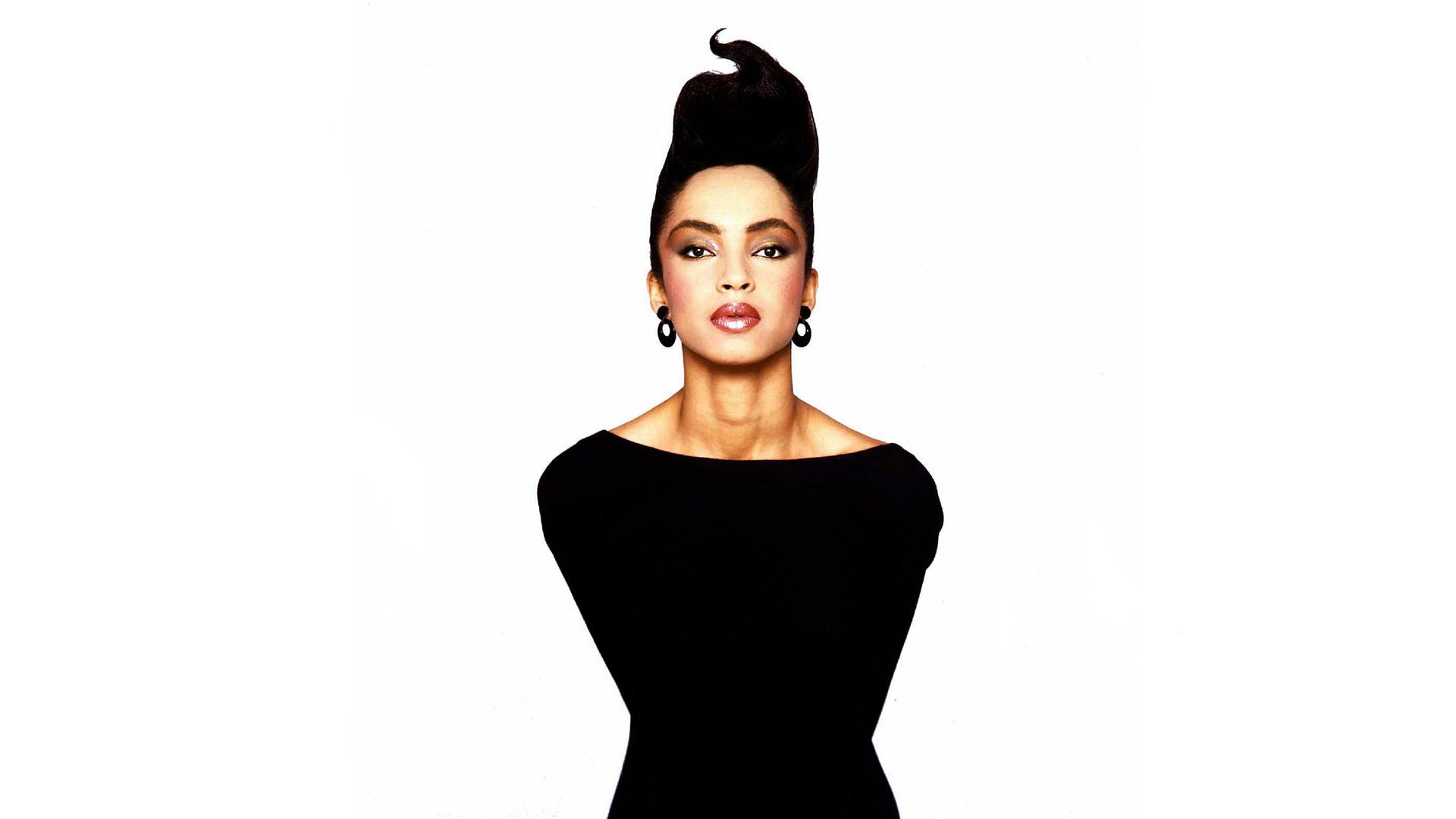 Die Sängerin Sade steht in ganz in schwarz gekleidet und die dunklen Haaren nach oben frisiert vor einem weißen Hintergrund. Sie lehnt sich vor und guckt in die Kamera.