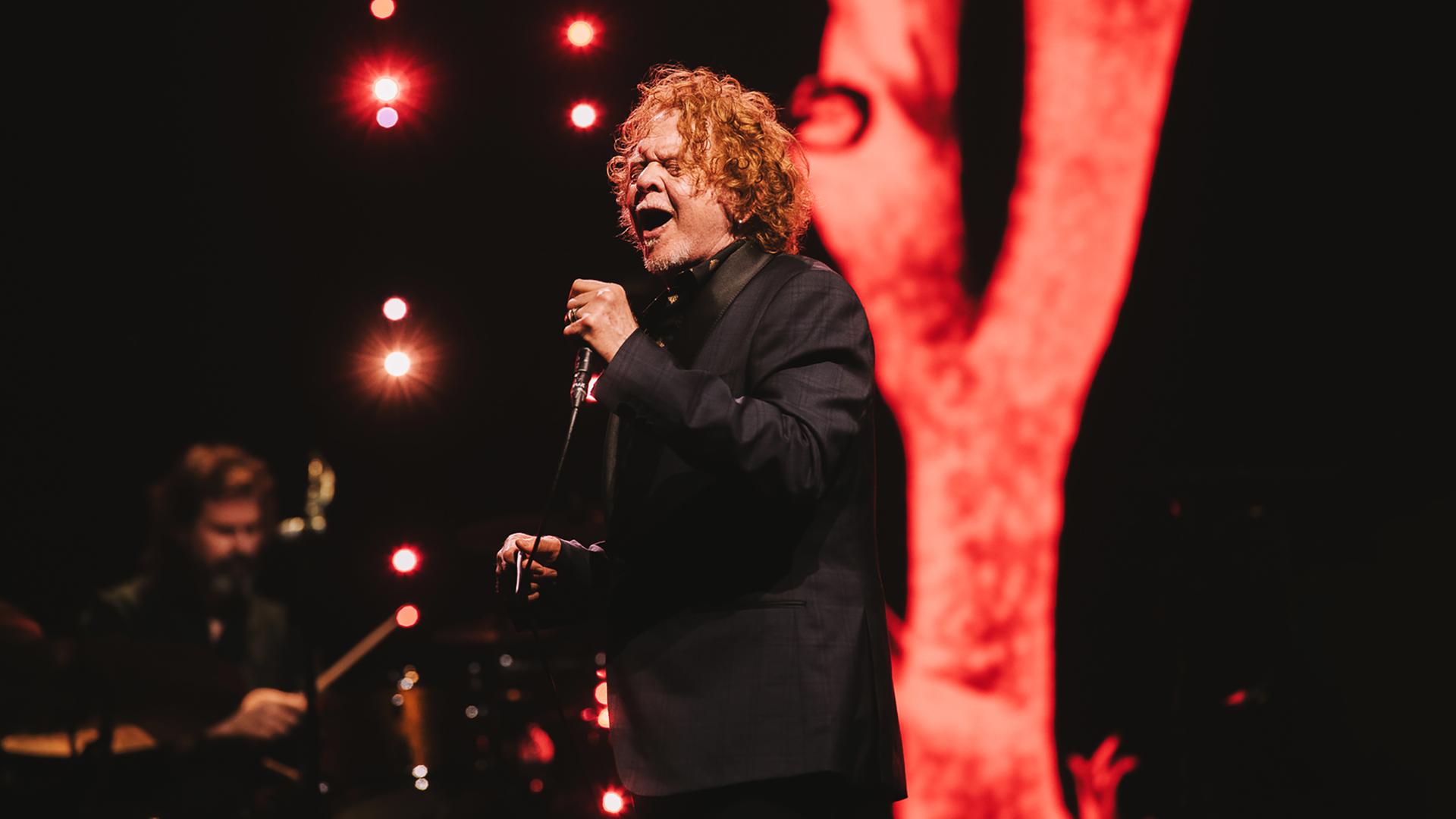 Mick Hucknall steht im schwarzen Anzug auf der Bühne und hält das Mikrofon mit beiden Händen und sing hinein.