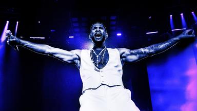 Usher steht mit offenen Armen und Mikro in der Hand auf der Bühne und singt.