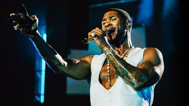 Usher steht auf der Bühne und singt ins Mikro während er mit der anderen Hand in den Zuschauerraum zeigt.