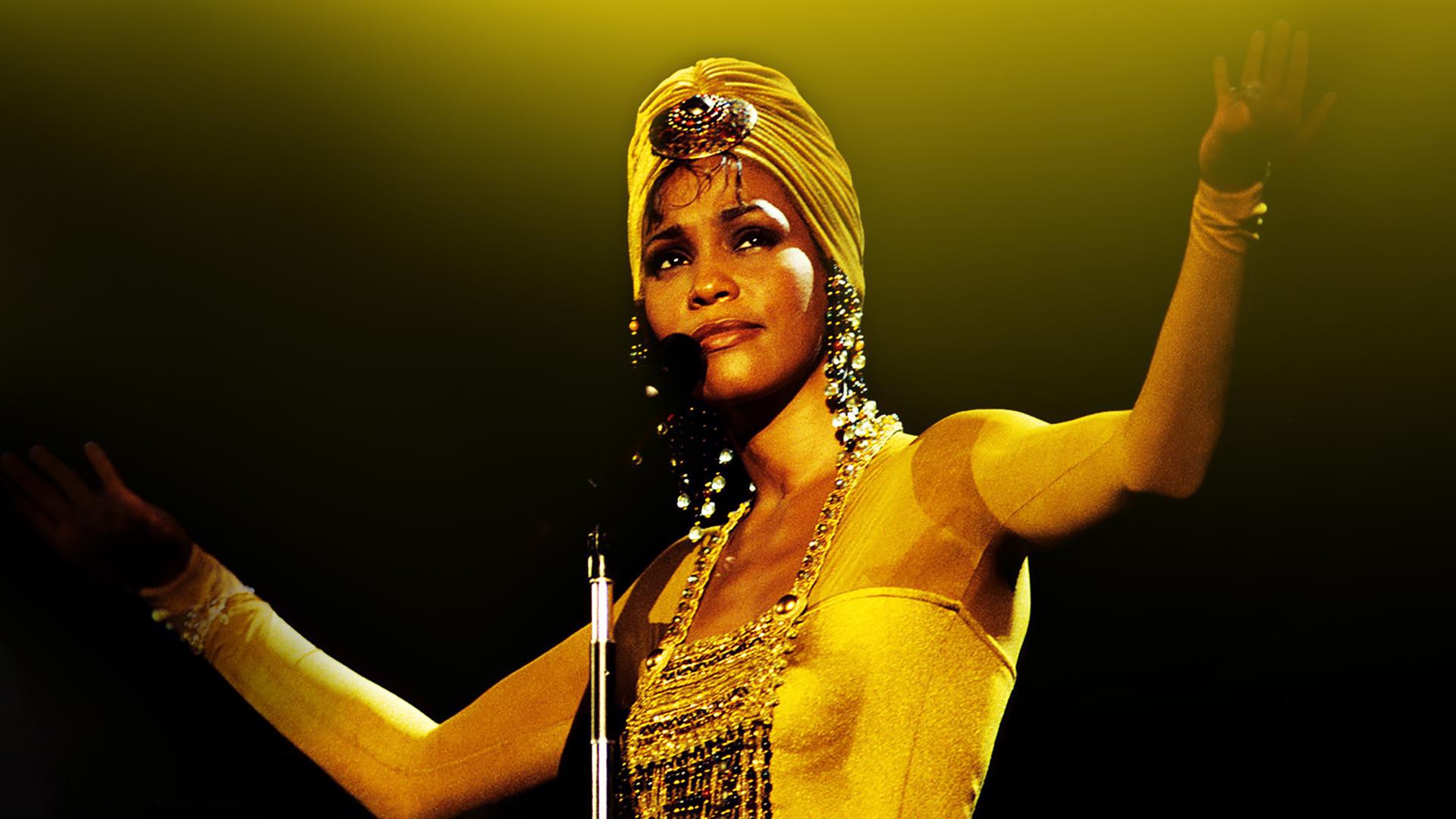 Whitney Houston steht vor einem Mikrofon, die Arme offen erhoben in der Luft. Die Haare trägt sie in einem Turban und sie trägt glitzernde Ohrringe und Oberteil.