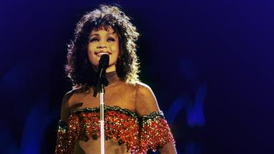 Die junge Whitney Houston steht lächelnd vor einem Mikrofon auf einer Bühne. 