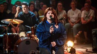 Loi sitzt auf einem Hocker und singt ins Mikrofon. Hinter ihr ist der Schlagzeuger mit Schlagzeug und Publikum zu erkennen.