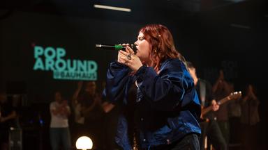 Loi singt ins Mirkofon. Hinter ist Publikum und ein der Pop Around Bauhaus Schriftzug zu sehen.