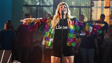 MIA steht mit offenen Armen und in regenbogenfarbener Glitzerfransenjacke und T-Shirt mit der Aufschrift "Liebe" auf der Bühne und singt ins Mikrofon. 