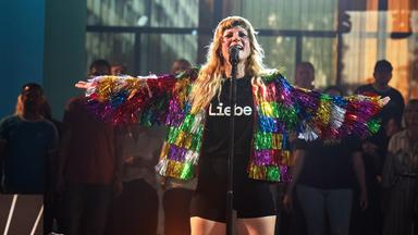 MIA steht mit offenen Armen und in regenbogenfarbener Glitzerfransenjacke und T-Shirt mit der Aufschrift "Liebe" auf der Bühne und singt ins Mikrofon. 