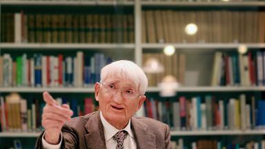 Jürgen Habermas sitzt vor einem Bücherregal und zeigt mit dem Finger an der Kamera vorbei.