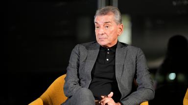 Ein älterer Mann im dunkelgrauen Anzug sitzt vor einem dunklen Hintergrund auf einem schwarzen Sessel.