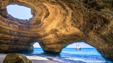 Eine Felshöhle, die eine halb im Meer gelegene Kuppel bildet und mehrere Eingänge besitzt. Im Hintergrund ein Schiff