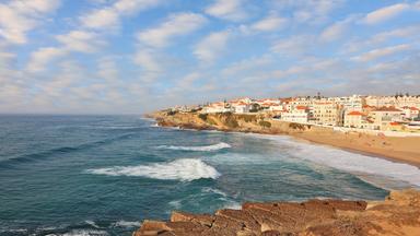 Blick von der Küste aus auf das Meer und die kleine Sadt Sintra mit Strand und vielen weißen Häusern und orangenen Dächern.