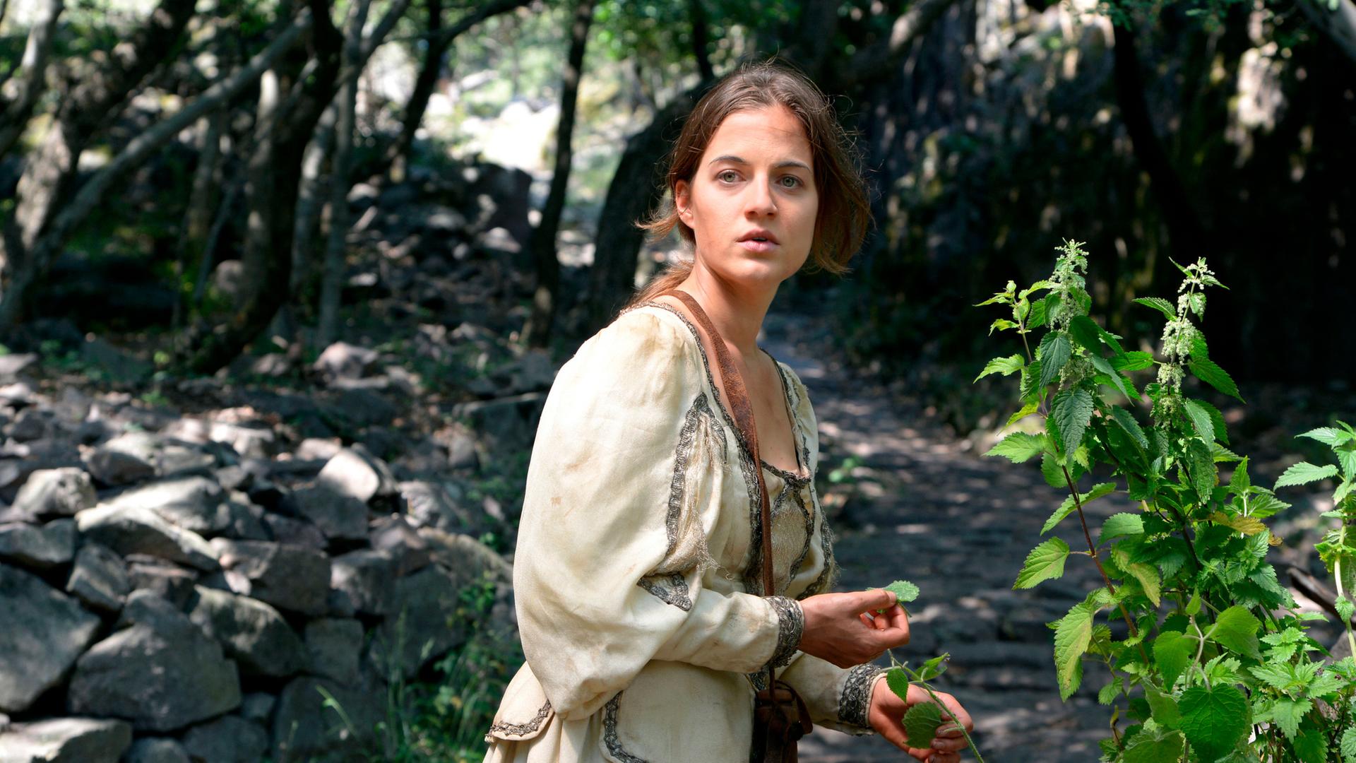 Eine Frau in einem dreckigen Kleid pflückt auf einem steinigen Weg Blätter von einem Brennesselstrauch.