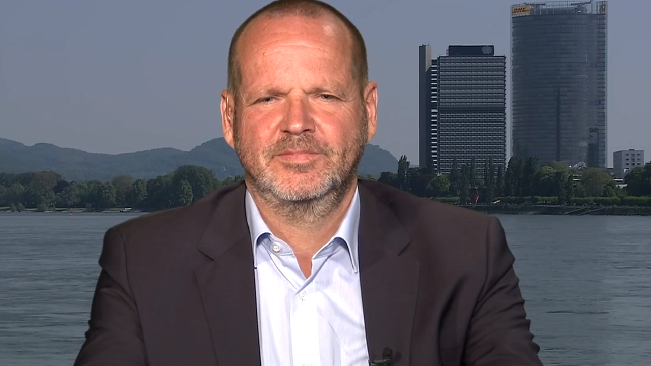 Mann im Anzug vor Fluss mit Hochhäusern im Hintergrund.