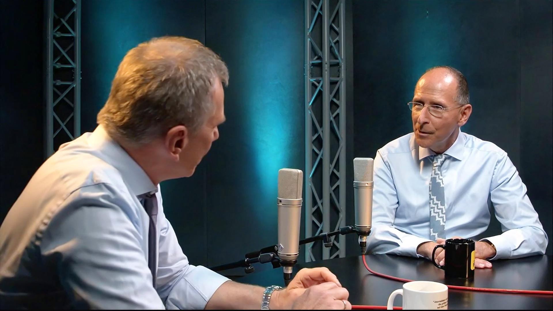 Die Abbildung zeigt zwei Männer, die in einem Studio miteinander sprechen. Es ist eine Multimedia-Sendung zu sehen, die sich mit dem politischen System in Österreich beschäftigt.    Im Vordergrund stehen zwei Mikrofone auf einem schwarzen Tisch. Der Mann links hat kurzes, graues Haar und trägt ein helles Hemd sowie eine Krawatte in dunklerer Farbe. Er schaut den anderen Mann an, dessen Gesicht von der Seite sichtbar ist.   Der Mann rechts hat eine Glatze und trägt eine Brille. Er hat ein hellblaues Hemd an und eine gemusterte Krawatte. Vor ihm steht eine schwarze Tasse. Die Wände des Studios sind dunkel und mit blauen Lichtakzenten beleuchtet, die im Hintergrund sichtbar sind.   Im Bild ist eine entspannte Atmosphäre zu erkennen, die auf ein informatives Gespräch hindeutet.
