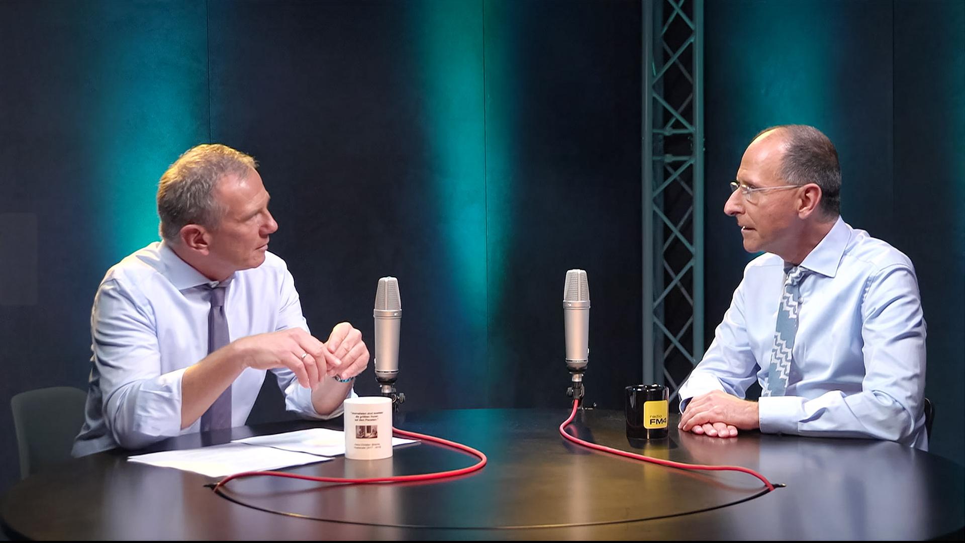 Das Bild zeigt zwei Männer, die an einem runden Tisch in einem Studio sitzen. Die Männer sind in formeller Kleidung gekleidet: der linke trägt ein weißes Hemd mit einem lila Krawatte, und der rechte ein hellblaues Hemd mit einem gemusterten, hellblauen Schlips.   Vor ihnen befinden sich zwei Mikrofone und ein schwarzer Becher mit dem Aufdruck „FM4“. Auf dem Tisch liegt ein Blatt Papier. Die Hintergrundbeleuchtung des Studios ist in sanften Blautönen gehalten. Der Raum hat eine minimalistische Gestaltung und vermittelt eine professionelle Atmosphäre.   Im Kontext des Bildes wird auf eine Sendung hingewiesen, die sich mit dem politischen System in Österreich beschäftigt.