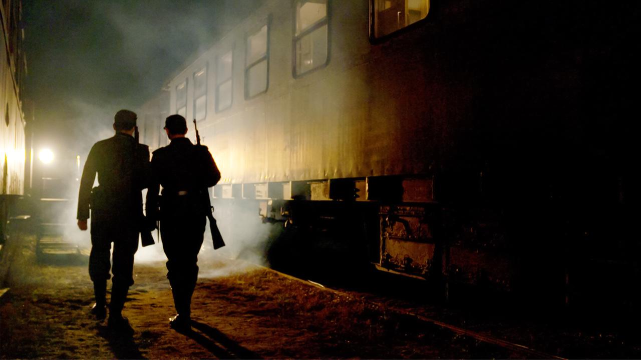 Das Bild zeigt eine nachgestellte Szene, die in einem schmalen, rautenförmigen Raum spielt, der von schummrigem Licht durchflutet wird. Im Vordergrund sind zwei Personen zu sehen, die nebeneinander gehen. Beide tragen dunkle, historische Uniformen, die an die Zeit des Nationalsozialismus erinnern. Eine der Personen hält ein Gewehr in der Hand.   Im Hintergrund ist ein Zug zu erkennen, der noch im Dämmerlicht steht. Die Wagen des Zuges sind dunkel und wirken altmodisch, mit wenigen erkennbaren Fenstern. Um den Zug herum schwebt leichter Nebel, was zu einer düsteren Stimmung beiträgt. Auf der linken Seite des Bildes gibt es eine Lichtquelle, vermutlich eine Lampe, die einen sanften Lichtstrahl auf den Boden wirft. Die gesamte Szenerie vermittelt ein Gefühl von Geheimnis und Anspannung, angedeutet durch die dämpfende Atmosphäre und die historische Kulisse.