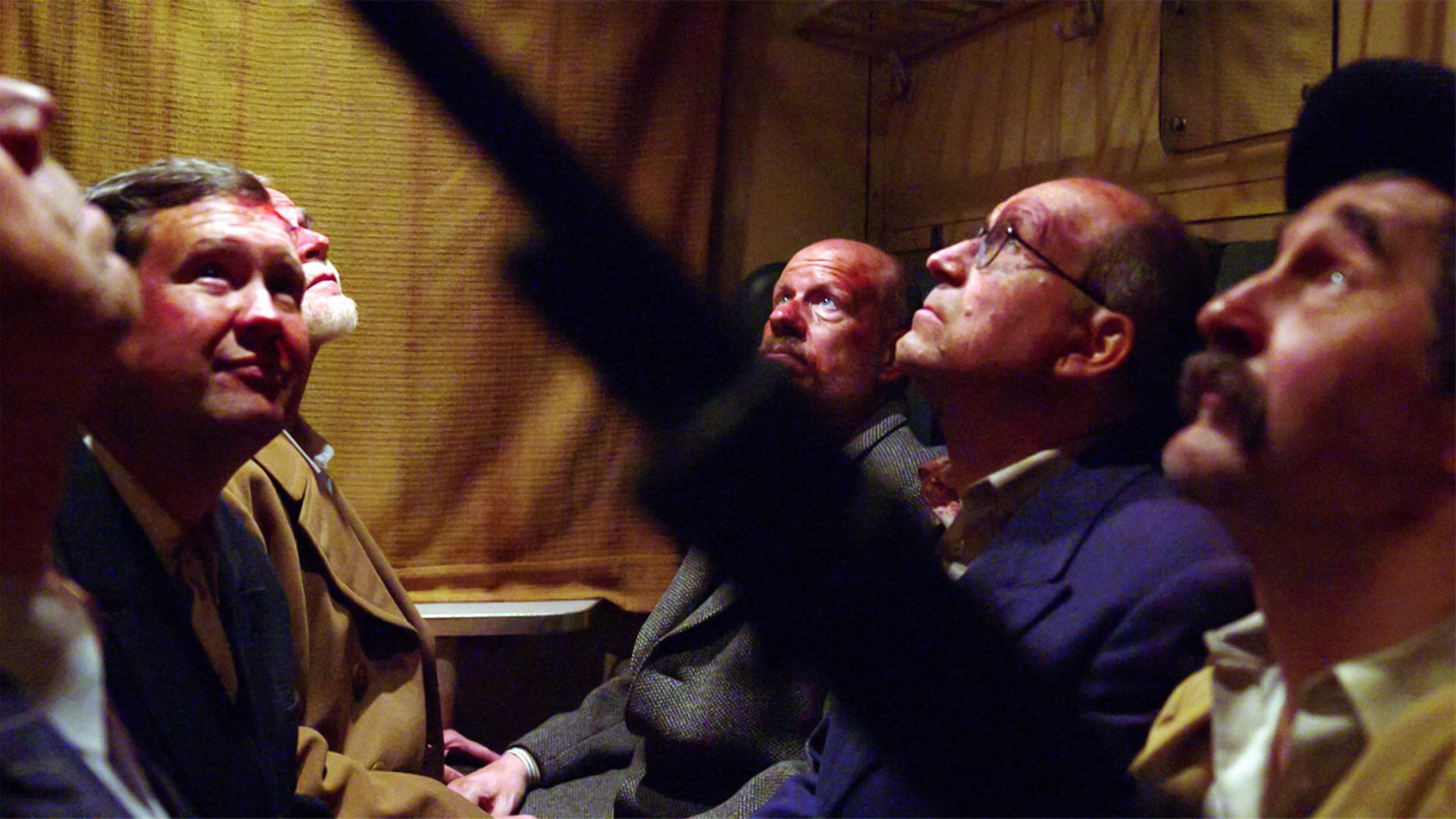 Das Bild zeigt eine nachgestellte Szene aus dem Film "Der Prominententransport 1938 - Mythos der Lagerstraße". In einem geschlossenen Raum, vermutlich einem Zugabteil, sitzen sechs Männer, die nachdenklich nach oben schauen.   Die Wände des Abteils sind mit einer hellbraunen, strukturierten Stoffverkleidung bedeckt. Die Männer tragen verschiedene historische Anzüge aus der Zeit der ersten Republik in Österreich, was auf die Jahre vor dem Zweiten Weltkrieg hinweist. Ihre Gesichtsausdrücke zeigen Besorgnis und Nachdenklichkeit. Die Beleuchtung ist schwach und erzeugt eine ernste Stimmung.  Im Hintergrund sind nur schwache Umrisse von weiteren Sitzplätzen und einigen Gegenständen erkennbar. Es gibt keine Fenster, die Sicht nach außen ist also nicht möglich.   Das Bild vermittelt eine Atmosphäre der Anspannung und des Unbehagens, passend zur Thematik der Verfolgung und Unterdrückung in der Zeit des Nationalsozialismus.