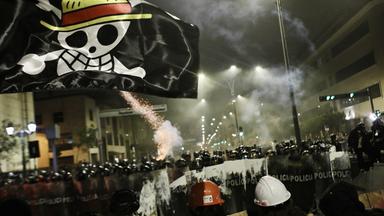 Demonstration bei Nacht: Polizisten mit Schilden sperren Demonstranten den Weg ab. Die Nacht wird von Straßenlampen und Feuerwerkskörpern erleuchtet. Über allem schwebt eine scharze Flagge mit einer Skelett-Figur, die einen Strohhut trägt