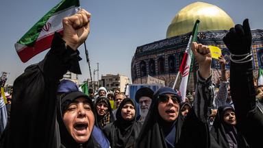 Frau ruft mit erhobener Faust vor iranischer Flagge, im Hintergrund eine Moschee.