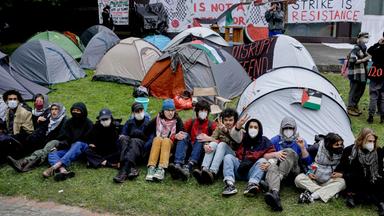 Eine Reihe junger Menschen mit Gesichtsmasken sitzen nebeneinander mit verschränkten Armen auf dem Rasen. Hinter ihnen sind Zelte aufgebaut und Plakate mit Slogans gegen den Krieg in Gaza aufgestellt