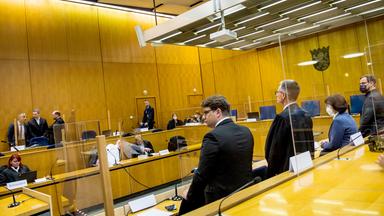 Mit Holz vertäfelter Gerichtssaal, an dessen Längsseiten jeweils zwei Reihen Podeste mit Stühlen angebracht sind, auf denen vereinzelt Menschen sitzen und stehen.