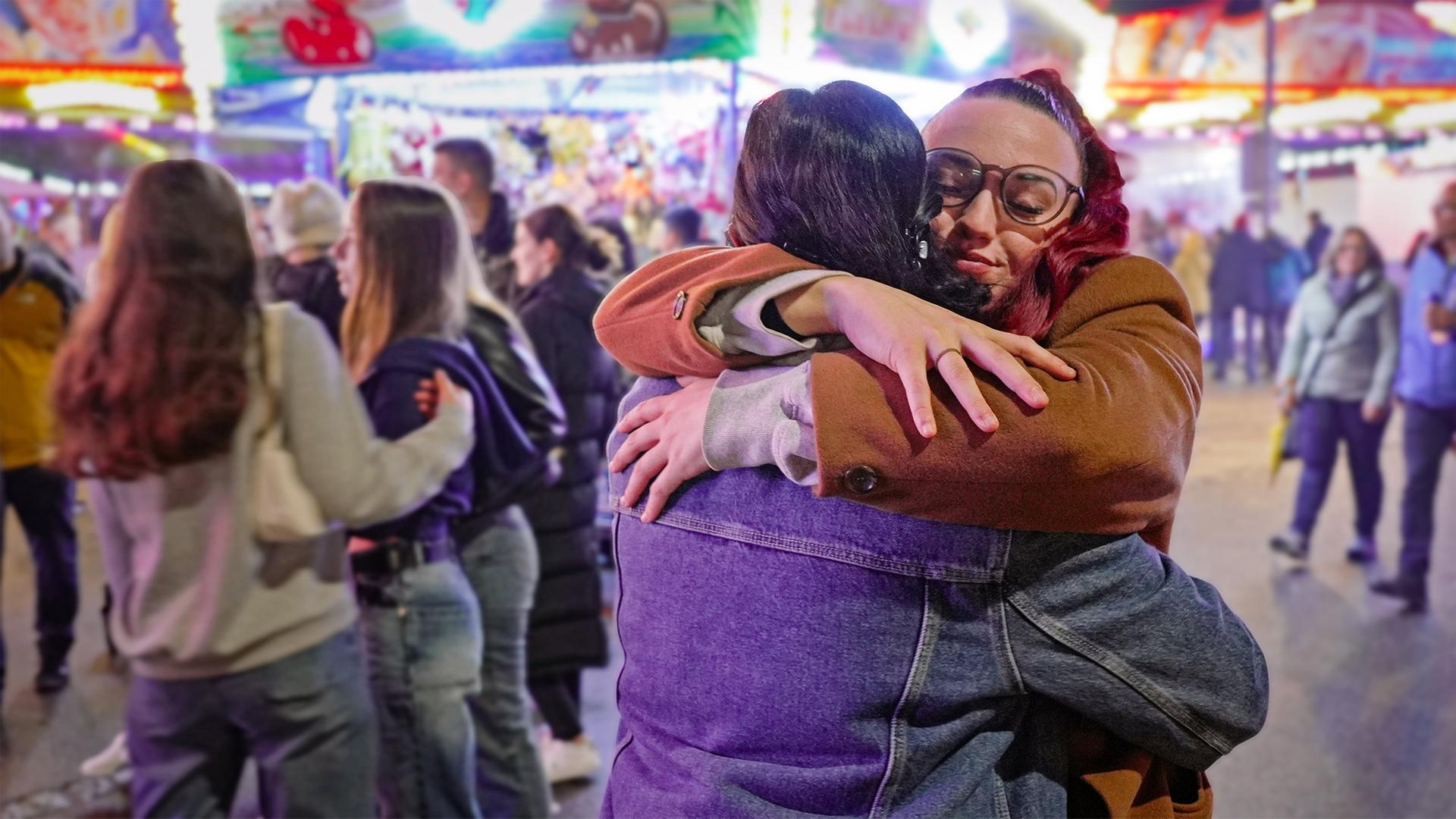 Zwei junge Frauen umarmen sich innig auf einem hell erleuchteten Jahrmarkt  im Hintergrund tummeln sich viele Menschen zwischen bunten Lichtern und Fahrgeschäften. Tosca und Angelina zeigen emotionale Verbundenheit.