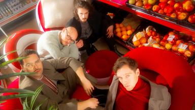 Zu sehen sind vier Männer, die in einem Supermarkt auf Sesseln im Gang sitzen. Hinter ihnen ist das Obst und Gemüse.