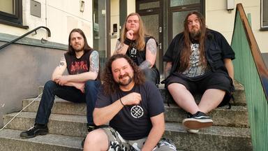 Vier Männer mit langen Haaren sitzen auf den Stufen eines Hauseingangs und posieren für die Kamera.